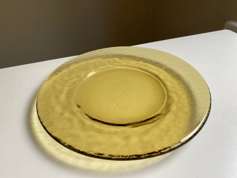 May include: A large, amber-colored glass plate with a textured surface. The plate has a slightly raised rim and a smooth, circular center.