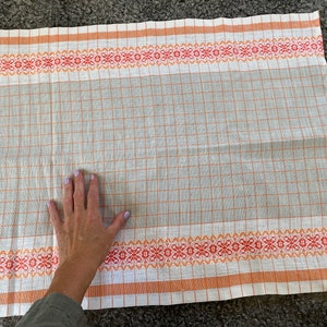 May include: A white kitchen towel with orange and red checkered pattern and a woven border. The towel is folded in half and laid flat on a grey surface.