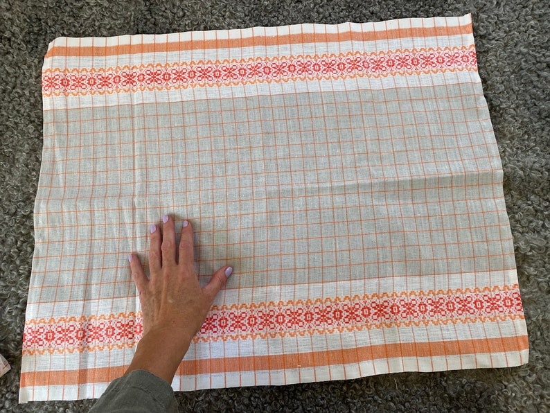 May include: A white kitchen towel with orange and red checkered pattern and a woven border. The towel is folded in half and laid flat on a grey surface.
