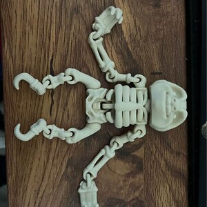 May include: A small, off-white articulated skeleton toy, lying on a wooden surface. The skeleton has a skull, rib cage, arms, and legs, all connected by joints. The toy is designed to be posed and is a popular novelty item.