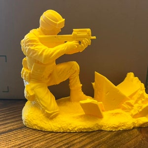 以下が含まれることがあります： 黄色い3Dプリントされた兵士のフィギュア。兵士はひざまずいてライフルを構えています。兵士はヘルメットとベストを着ています。フィギュアは、兵士の後ろに破壊された建物がある台座の上に置かれています。