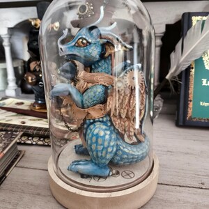 Op de afbeelding: Een blauw drakenbeeldje met gouden accenten, ingesloten in een glazen stolp. De draak heeft gedetailleerde schubben en vleugels en is gemonteerd op een houten voet. Het beeldje heeft een steampunk-esthetiek.