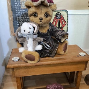 May include: A plush teddy bear wearing a burgundy hat and coat, holding a stuffed bunny. The bear and bunny are on a wooden desk. A chalkboard with French text is in the background. A decorative tin with a monkey is also visible.
