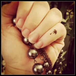 May include: A close-up of a person's finger with a small palm tree tattoo. The finger is adorned with a silver chain necklace with round beads.