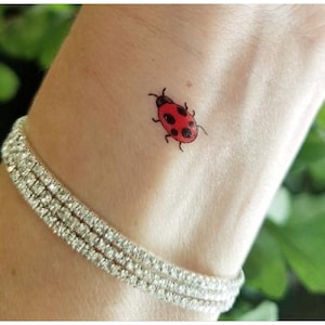 May include: A small red ladybug tattoo on a person's wrist. The tattoo is a simple design with black outlines and red spots.