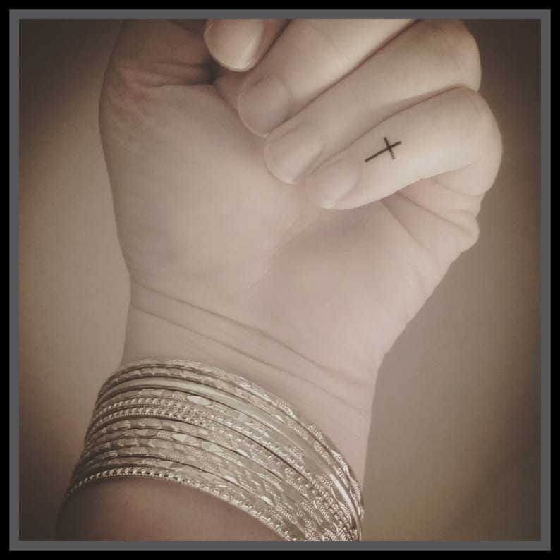 May include: A close-up of a hand with a small cross tattoo on the finger. The hand is wearing a stack of silver bangles.