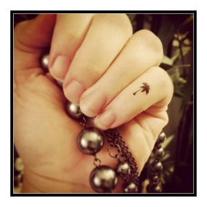 May include: A close-up of a person's finger with a small palm tree tattoo. The finger is adorned with a silver chain necklace with round beads.