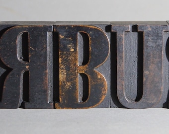Op de afbeelding: Een close-up van houten letterblokken die het woord "HOBBY" in hoofdletters spellen. De blokken zijn donkerbruin met een versleten, verweerde afwerking.