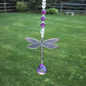 May include: A silver dragonfly suncatcher with a purple crystal teardrop hanging from it. The dragonfly is suspended from a chain with clear and purple beads.
