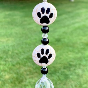 May include: A car charm featuring two white ceramic paw print beads with black paw prints, separated by black beads. A clear crystal hangs from the bottom.