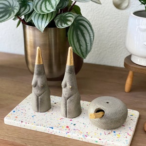 Peut inclure: Deux sculptures coniques grises avec des pointes dorées et de petits accents dorés, exposées sur un plateau en terrazzo. Un porte-encens rond gris avec un bâtonnet en bois est également sur le plateau. Une plante en pot est en arrière-plan.