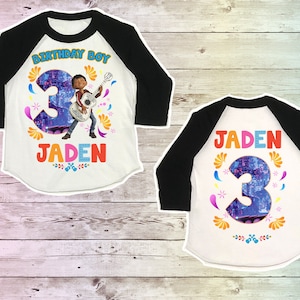 May include: A white and black baseball style shirt with a colorful design featuring a cartoon character playing a guitar and the number 3. The text "Birthday Boy" and "Jaden" are printed on the shirt.