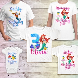 Puede incluir: Conjunto de cinco camisetas blancas con tem&aacute;tica de sirena. Las camisetas tienen diferentes textos e im&aacute;genes, incluyendo "Pap&aacute; de la ni&ntilde;a del cumplea&ntilde;os", "Mam&aacute; de la ni&ntilde;a del cumplea&ntilde;os", "Hermano de la ni&ntilde;a del cumplea&ntilde;os", "Hermana de la ni&ntilde;a del cumplea&ntilde;os" y una camiseta con el n&uacute;mero 3 y el nombre "Olivia".
