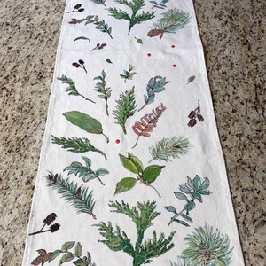 May include: White table runner with a botanical design. Features green and blue-green leaves, pine needles, and pine cones. Red dots are scattered throughout the design. The runner is rectangular and appears to be made of fabric.