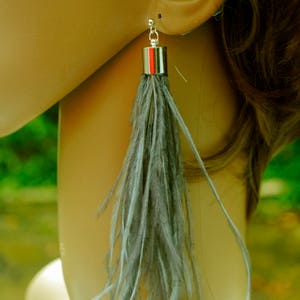 May include: A pair of long, gray feather earrings with silver metal accents. The earrings are dangling from the earlobes of a mannequin.