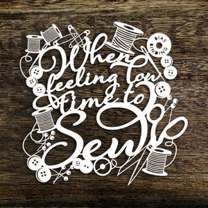 May include: White paper cut-out design on a wood background. The design features the text "When feeling low, time to sew" surrounded by sewing supplies like spools of thread, buttons, and safety pins.