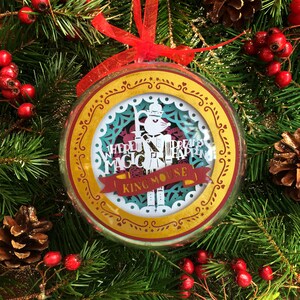 以下が含まれることがあります： 金と赤のクリスマスオーナメント。紙で切り抜かれたくるみ割り人形のデザインと「Where dreams happen, magic is King Mouse!」というテキストが書かれています。