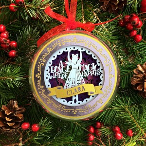 以下が含まれることがあります： 金色の縁取りとキラキラ光る紫色の背景を持つ丸いガラス製のクリスマスオーナメント。オーナメントには、「A Place Where Magic Happens」という文字と「Clara」という名前が書かれた金色のリボンが付いた、白い紙で切り抜かれたバレリーナのシルエットが描かれている。