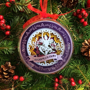 以下が含まれることがあります： 白い紙で切り抜かれた妖精のデザインが施された、紫と金のクリスマスオーナメント。妖精の周りに「A Land of Magic Sweets Sugar Plum Fairy」という文字が書かれています。