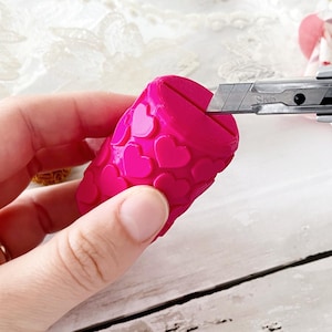 Op de afbeelding: Een felroze cilindervormig object met een herhalend reliëfpatroon van hartjes. Het object heeft een horizontale gleuf aan de bovenzijde en een zilverkleurig stanleymesje is in de gleuf geplaatst om de diepte en de werking ervan te demonstreren.