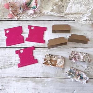 Può includere: Una collezione di materiali per l'artigianato tra cui etichette fustellate rosa acceso, linguette di cartoncino marrone piegate ed etichette decorative con pizzo e abbellimenti floreali. Disposti su una superficie di legno bianco.