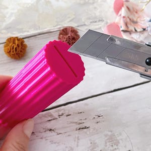 May include: A bright pink, cylindrical, fluted plastic object with a narrow vertical slit on the top surface. A silver metal utility knife with a sharp blade is positioned near the edge of the pink object, which has a textured, ridged exterior.