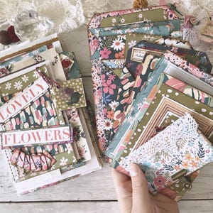 Könnte beinhalten: Zwei handgefertigte Junk Journals mit floralen und abstrakten Mustern, Spitzenakzenten und geschichteten Kartonelementen. Das linke Journal zeigt den Text "LOVELY FLOWERS" auf dem Einband, während das rechte Journal mehrere Einstecktaschen enthält.