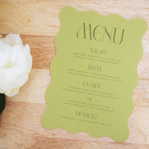 Puede incluir: Una tarjeta de men&uacute; verde claro con un borde ondulado, con la palabra "MENU" en la parte superior. El men&uacute; enumera opciones de ensalada, pasta, entrante y postre. Una flor de peon&iacute;a blanca con hojas verdes est&aacute; en el lado izquierdo, sobre un fondo de madera.