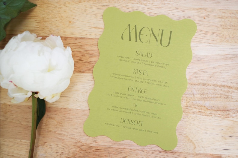 Puede incluir: Una tarjeta de men&uacute; verde claro con un borde ondulado, con la palabra "MENU" en la parte superior. El men&uacute; enumera opciones de ensalada, pasta, entrante y postre. Una flor de peon&iacute;a blanca con hojas verdes est&aacute; en el lado izquierdo, sobre un fondo de madera.
