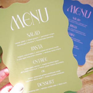 Puede incluir: Dos tarjetas de men&uacute; con borde ondulado. Una es de color verde claro con texto blanco, que enumera opciones de ensalada, pasta, entrante y postre. La otra es azul con texto rosa, que muestra la palabra "MENU". Se ven acentos florales.