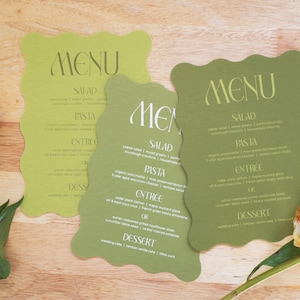 Puede incluir: Tres tarjetas de men&uacute; de boda verdes con un borde ondulado, con la palabra "MENU" en la parte superior. El men&uacute; incluye secciones para ensalada, pasta, entrante y postre. Una peon&iacute;a blanca y rosa est&aacute; en primer plano.