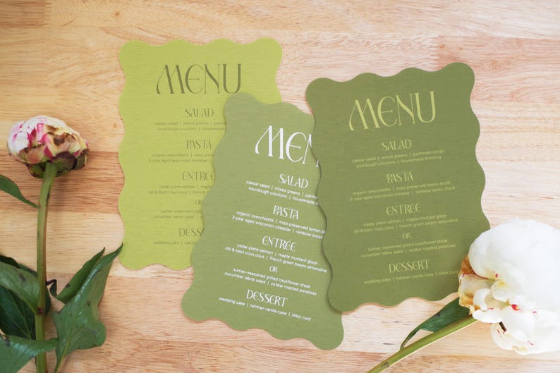 Puede incluir: Tres tarjetas de men&uacute; de boda verdes con un borde ondulado, con la palabra "MENU" en la parte superior. El men&uacute; incluye secciones para ensalada, pasta, entrante y postre. Una peon&iacute;a blanca y rosa est&aacute; en primer plano.