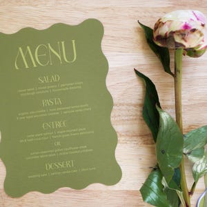 Puede incluir: Una tarjeta de men&uacute; verde salvia con un borde ondulado, con la palabra "MENU" en una fuente estilizada. El men&uacute; enumera opciones de ensalada, pasta, entrante y postre. Una flor de peon&iacute;a con p&eacute;talos rosas y blancos est&aacute; al lado del men&uacute;.