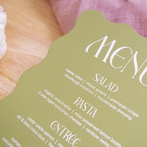 Puede incluir: Una tarjeta de men&uacute; verde claro con un borde ondulado, que muestra la palabra "MENU" en escritura blanca. El men&uacute; enumera ensaladas, pastas y platos principales. Una flor blanca y tela rosa est&aacute;n en el fondo.