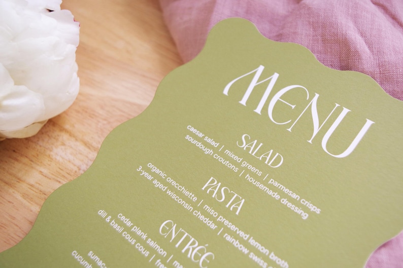 Puede incluir: Una tarjeta de men&uacute; verde claro con un borde ondulado, que muestra la palabra "MENU" en escritura blanca. El men&uacute; enumera ensaladas, pastas y platos principales. Una flor blanca y tela rosa est&aacute;n en el fondo.