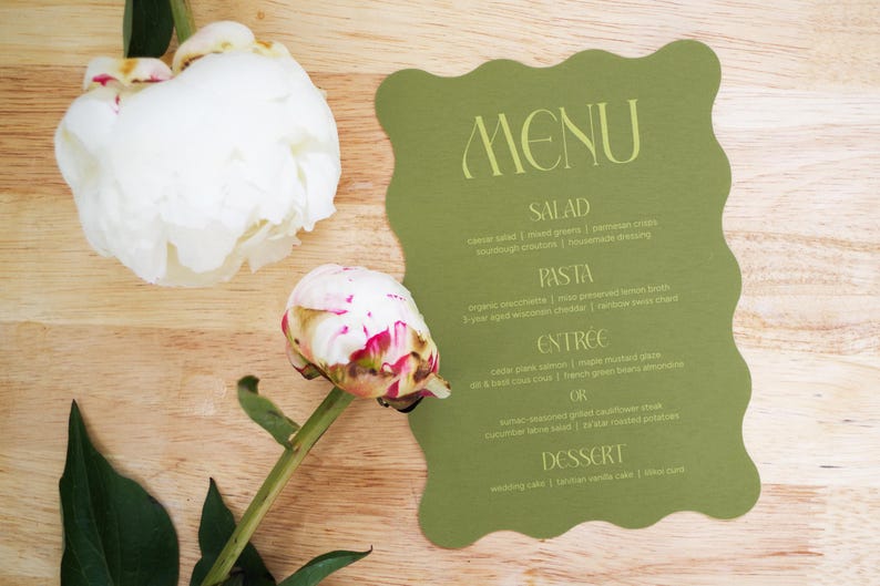 Puede incluir: Una tarjeta de men&uacute; verde con borde ondulado, con la palabra "MENU" en escritura elegante. El men&uacute; enumera ensalada, pasta, plato principal y postre. Dos peon&iacute;as blancas y rosas est&aacute;n sobre una superficie de madera.