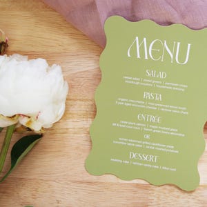Puede incluir: Una tarjeta de men&uacute; verde claro con un borde ondulado, con texto blanco. El men&uacute; enumera opciones de ensalada, pasta, entrante y postre. Una peon&iacute;a blanca y una tela rosa est&aacute;n en el fondo.