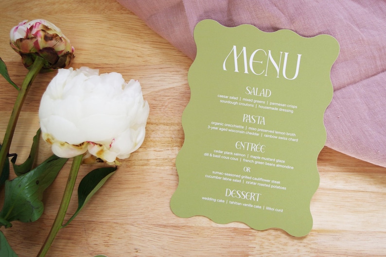 Puede incluir: Una tarjeta de men&uacute; verde claro con un borde ondulado, con texto blanco. El men&uacute; enumera opciones de ensalada, pasta, entrante y postre. Una peon&iacute;a blanca y una tela rosa est&aacute;n en el fondo.