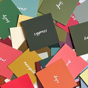 May include: A collection of colorful square cards in various shades of green, pink, orange, yellow, and more. Each card features a different word in white script, such as "cypress" and "kelp". The cards are scattered, creating a vibrant and artistic display.