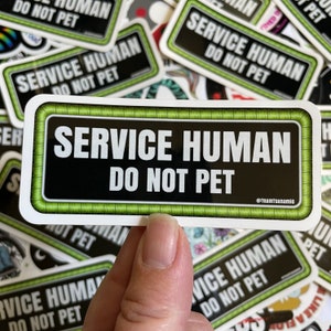 Op de afbeelding: Een zwarte en groene sticker met witte tekst die "SERVICE HUMAN DO NOT PET" luidt, met een groene rand en een kleine witte tekst onderaan die "@TeamTsunamis" luidt.