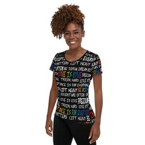 Pode incluir: Uma camiseta preta com uma estampa colorida das palavras "Love is Love" e outras mensagens positivas. A camiseta está sendo usada por uma pessoa com cabelo castanho e um sorriso.
