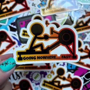May include: A white sticker with a black stick figure rowing a rowing machine. The sticker has a gradient background of orange, yellow, and red. The text "GOING NOWHERE..... FAST!" is printed on the sticker.