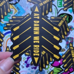 May include: Black and yellow sticker with the text "AT DAWN WE RIDE" in a stylized font. The sticker is shaped like an arrow pointing down and has a design of oars in the background.