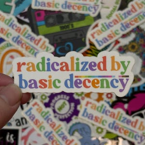 May include: A collection of stickers with the phrase "radicalized by basic decency" in a rainbow font. The stickers are white with a rectangular shape. Other stickers with similar text and designs are visible in the background.