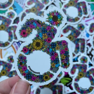 Pode incluir: Um adesivo branco com um design floral colorido em forma do símbolo Om. O adesivo tem um fundo branco e está rodeado por outros adesivos. O texto "@trevorsunami6" está na parte inferior do adesivo.