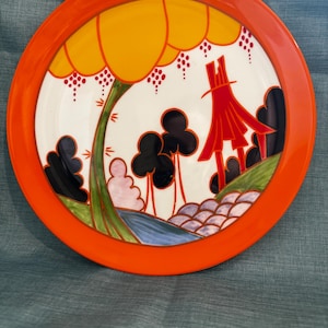 Puede incluir: Un plato redondo de cerámica con un borde ancho de color naranja sólido. El centro presenta una ilustración de paisaje estilizada con un gran árbol amarillo, una estructura roja tipo pagoda, árboles negros, un arroyo azul y formaciones rocosas moradas.