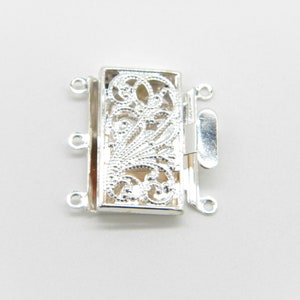 May include: Silver-toned jewelry clasp with an ornate, openwork design. The clasp features a rectangular shape with a decorative floral pattern and a secure closure mechanism. It has multiple loops for attaching to jewelry.