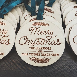 Puede incluir: Un conjunto de adornos redondos de madera con un dise&ntilde;o grabado con l&aacute;ser marr&oacute;n. Los adornos presentan el texto "Merry Christmas" y los nombres "The Claypools and Your Victory Ranch Crew".
