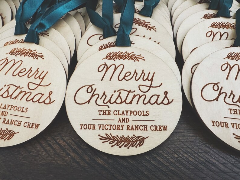 Puede incluir: Un conjunto de adornos redondos de madera con un dise&ntilde;o grabado con l&aacute;ser marr&oacute;n. Los adornos presentan el texto "Merry Christmas" y los nombres "The Claypools and Your Victory Ranch Crew".