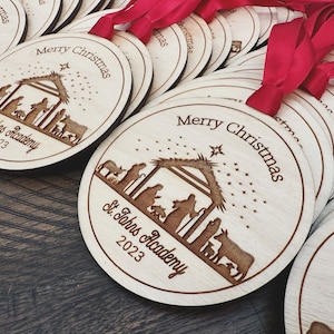 May include: A set of wooden Christmas ornaments with a nativity scene design. The ornaments are round and have a brown wood finish. The text "Merry Christmas" and "St. Johns Academy 2023" is engraved on the ornaments.